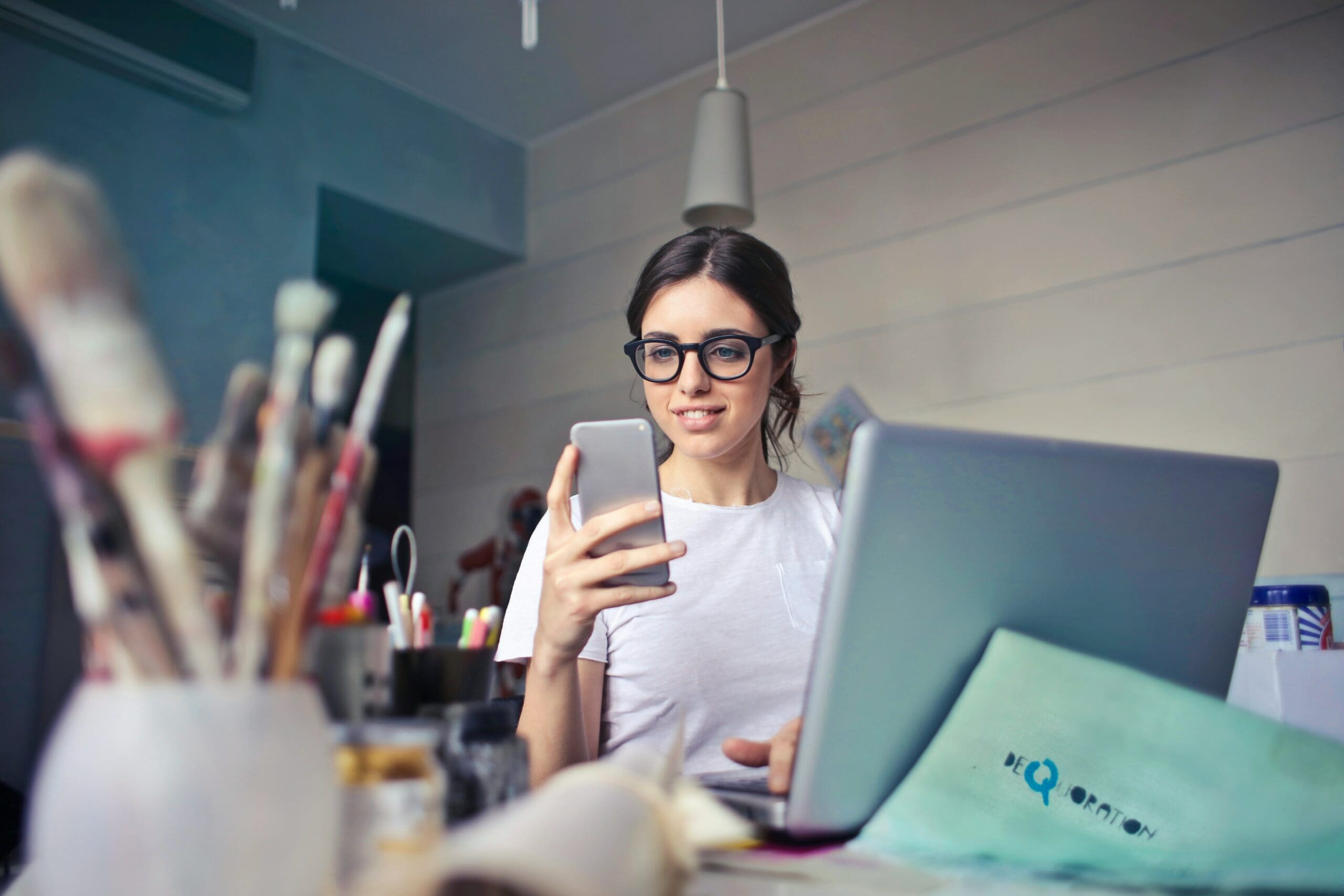 woman sits at an art desk with a laptop, looking at her phone