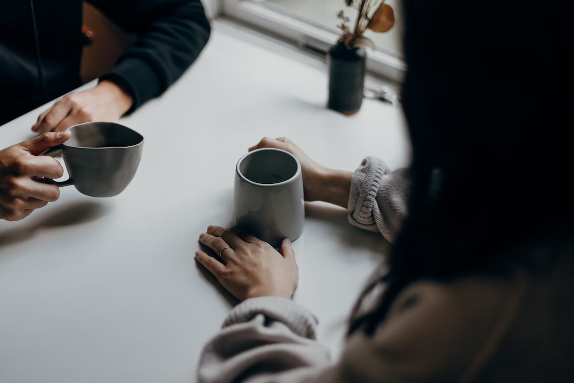 two people at a table talking with mugs in their hands