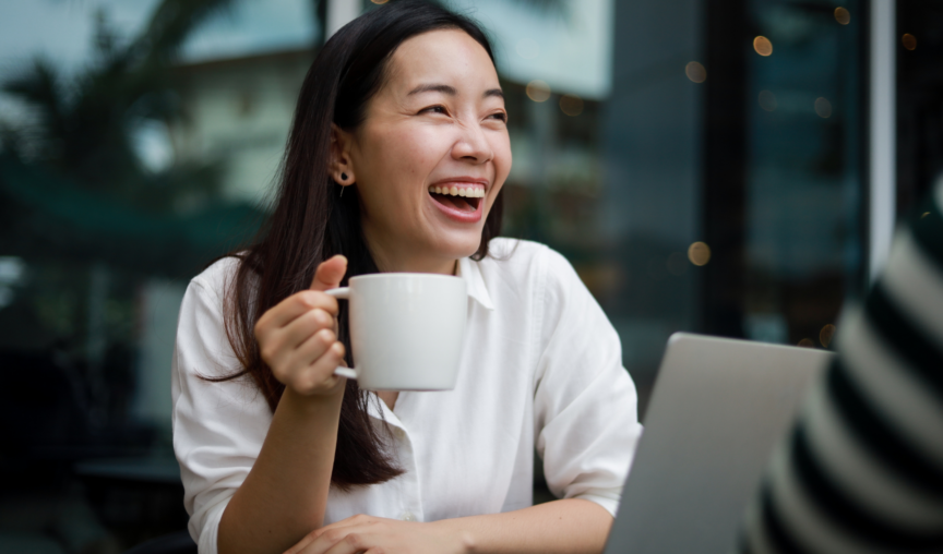 women holding a cup of coffee and smiling in front of her laptop while working