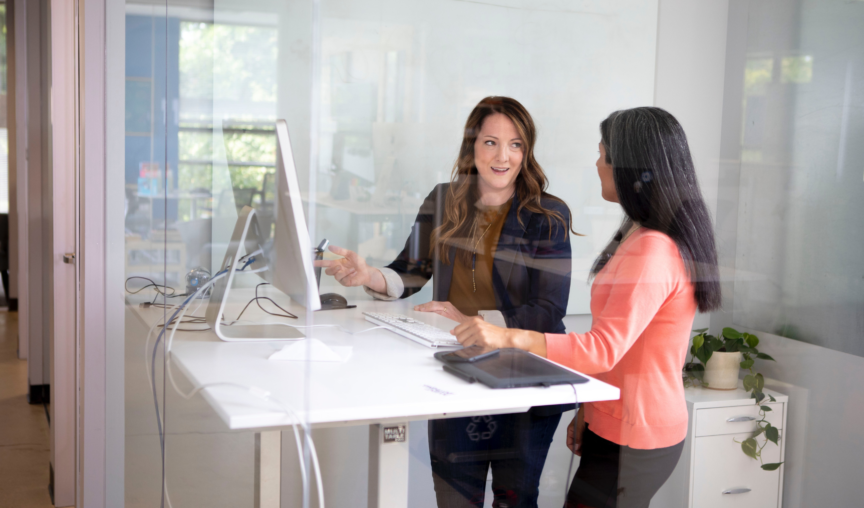 Two woman standing at a desk, talking in front of a computer