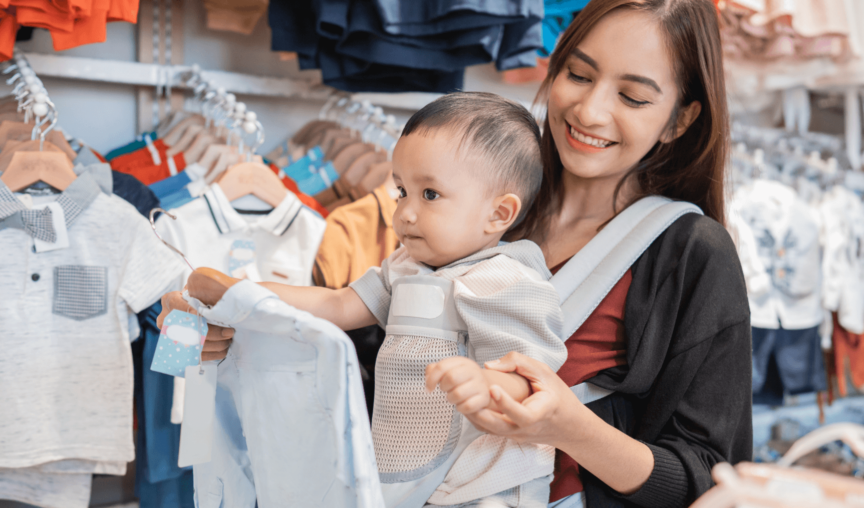 Mom shopping in a store while holding up a piece of baby's clothing in front of her kid in a carrier
