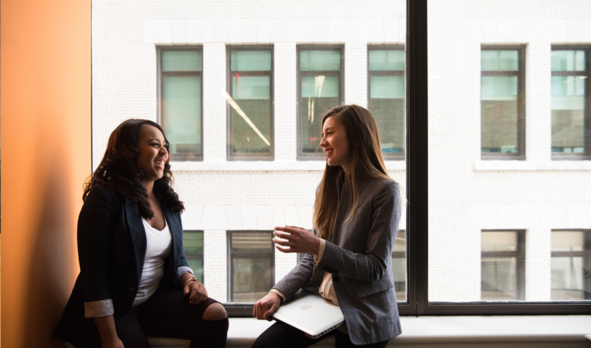 Two women of different ethnicities sitting together at an office window chatting