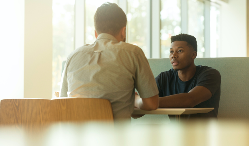 Two men sit at a table, looking serious