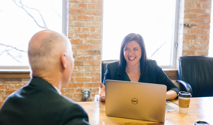 A woman smiles over a laptop at a man facing away from the camera