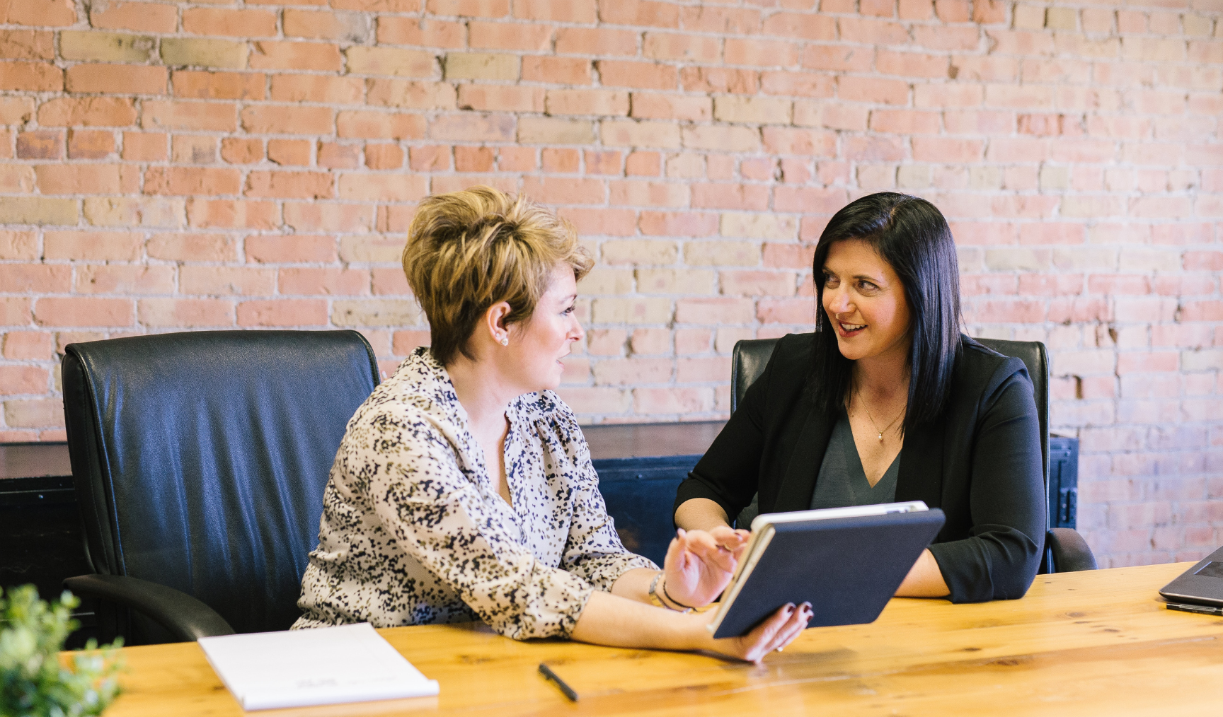 Two woman sit at a large table with a tablet held between them, in deep discussion