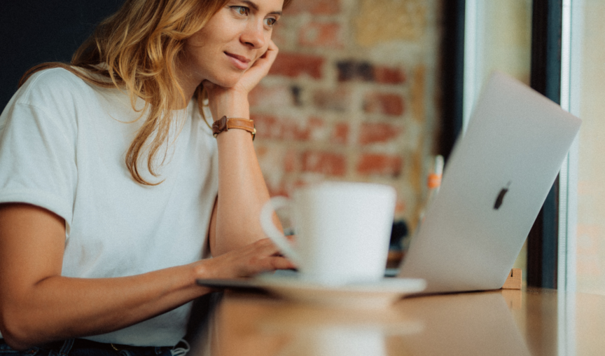 A woman sits in a cafe, looking at her laptop with a mug of coffee next to her