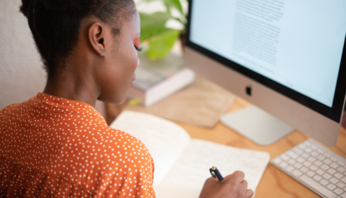 A woman writes notes in a notebook in front of a computer