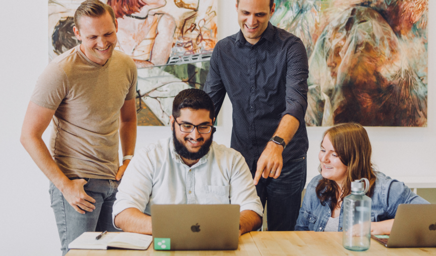 Man sits at a table on his laptop, while his colleagues surround him observing his work