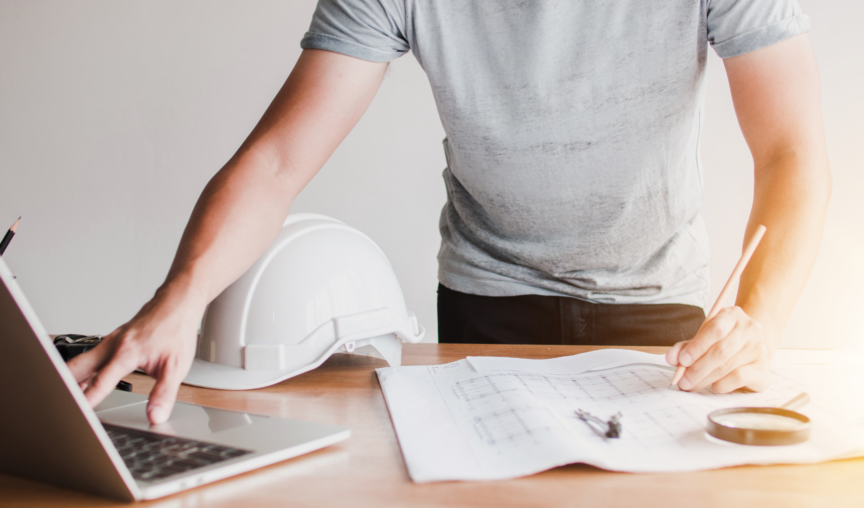 A man works at a desk, construction helmet on the desk and laptop there