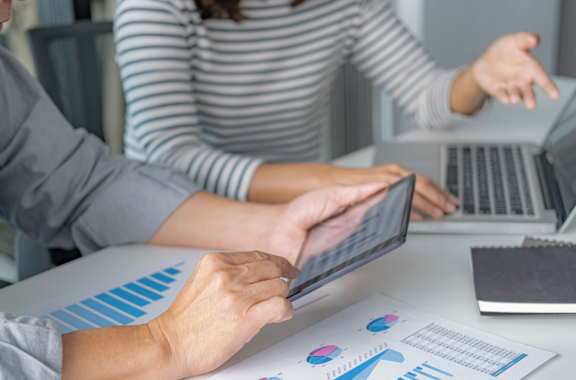 Two people sit at a desk, discussing something. Their torsos and arms are only visible - one has a laptop, the other a tablet