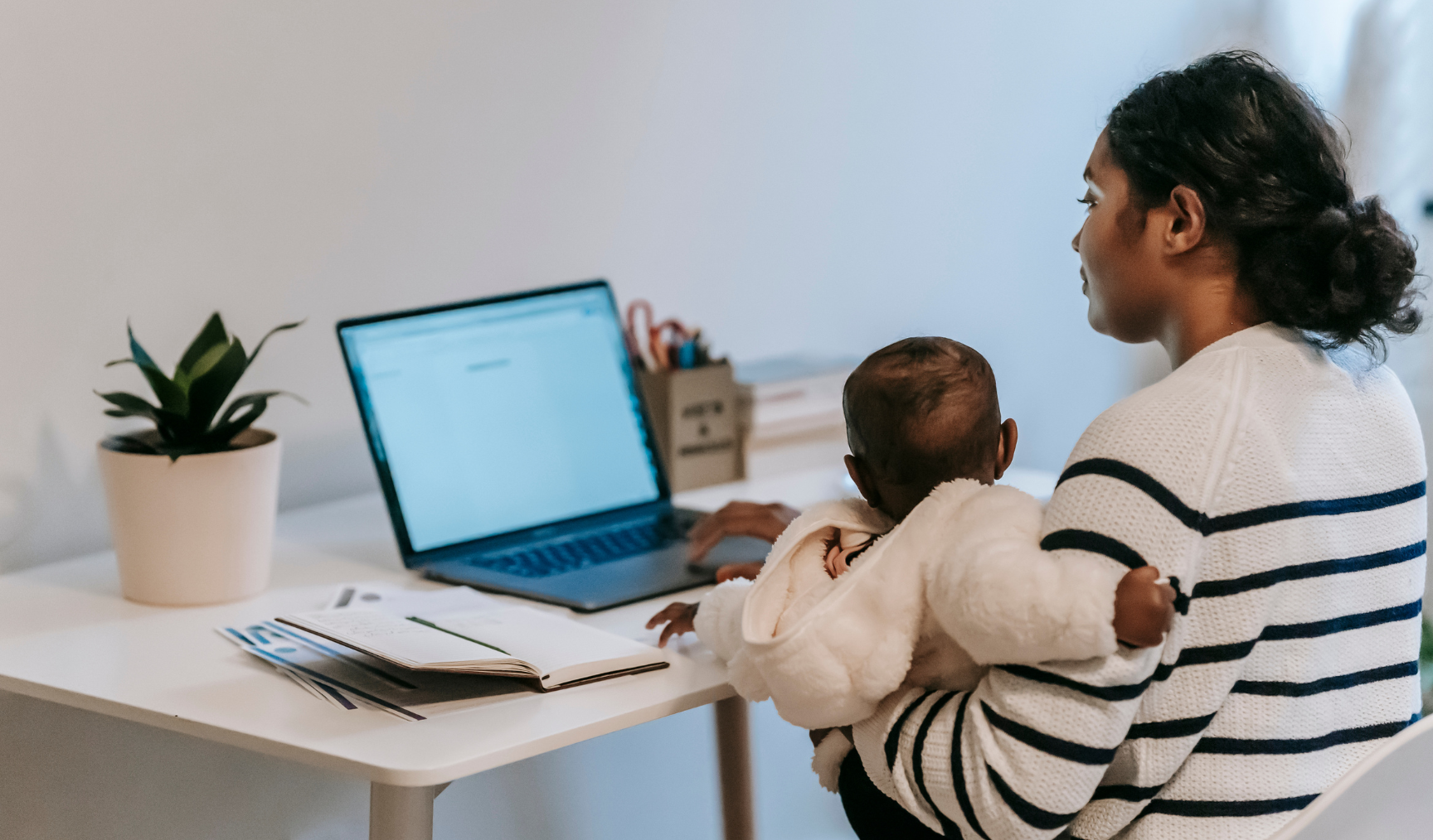 Woman sits at a laptop, with a baby in her arms
