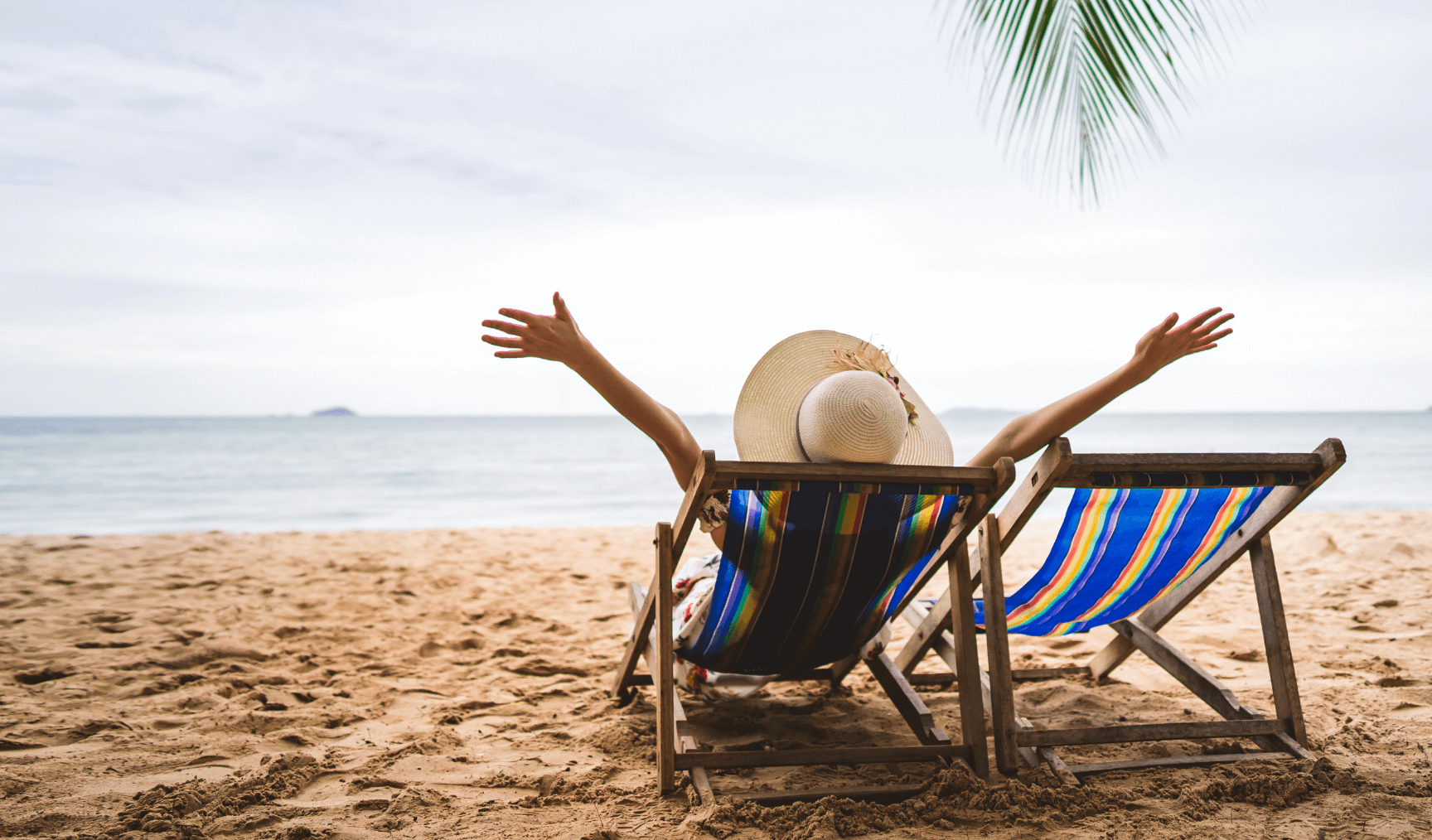 person in a sun hat sitting on a lounge chair on the beach with their arms outstretched