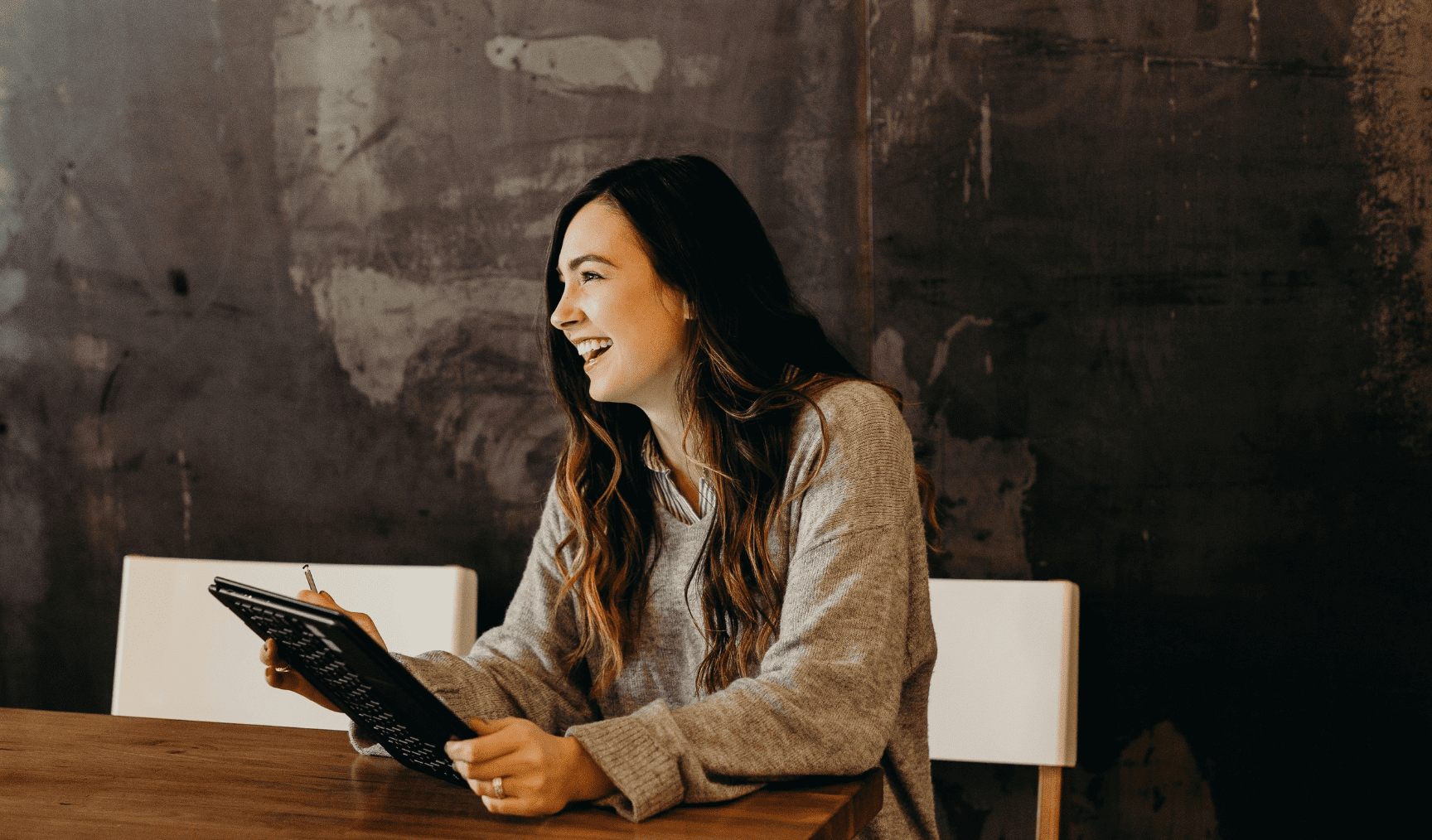 A woman sits at a table, tablet in hand, smiling