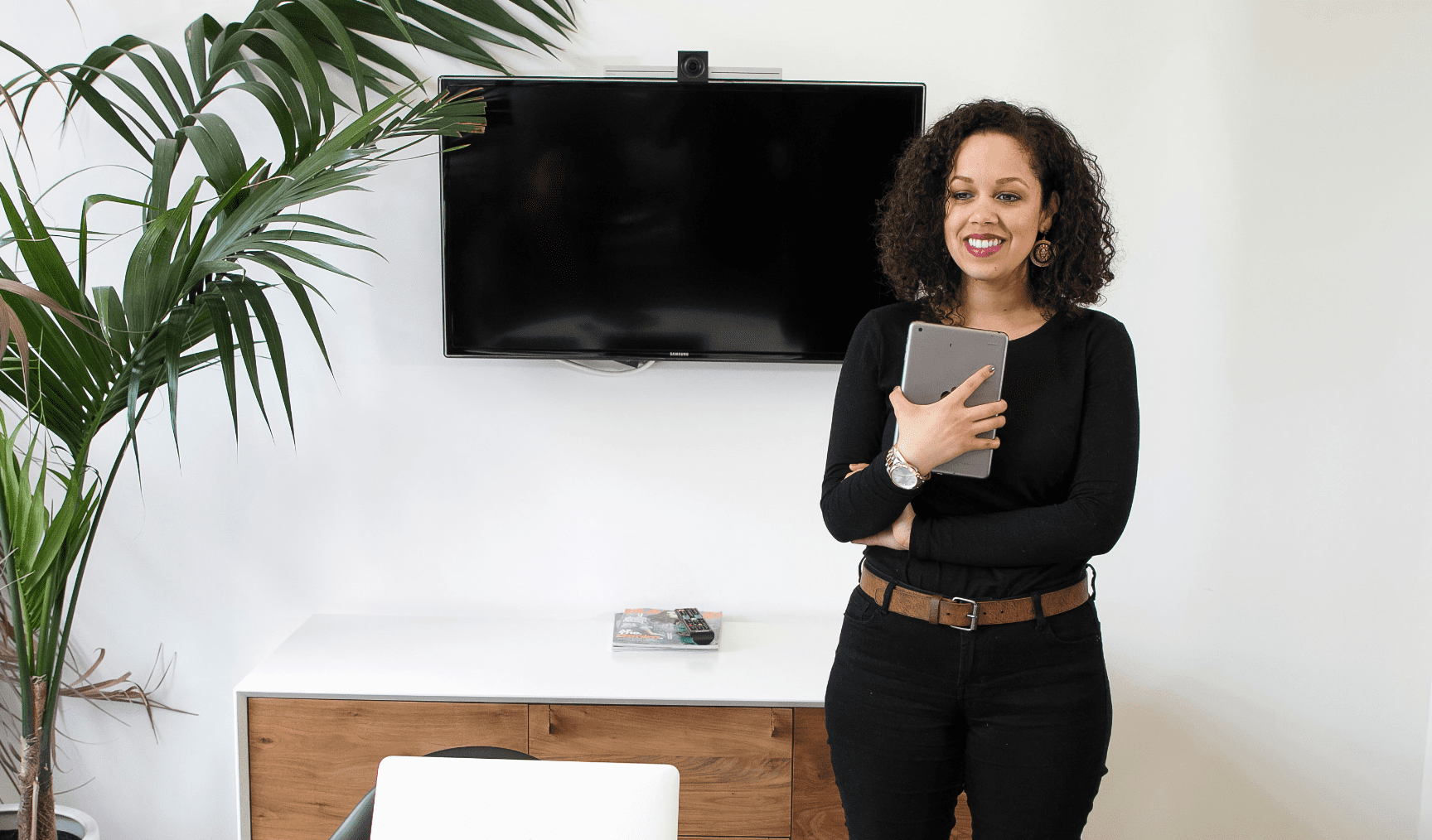 A woman stands at the head of a meeting room, tablet in hand, smiling