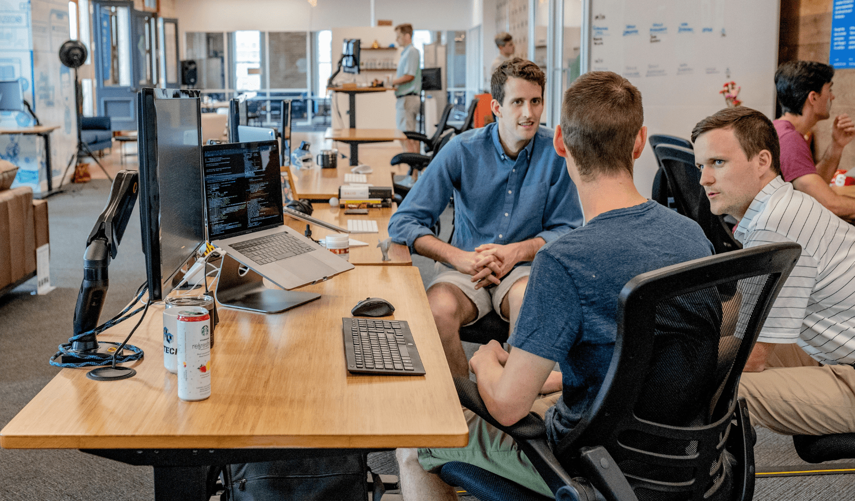 Three men sit at a desk in an office, chatting casually