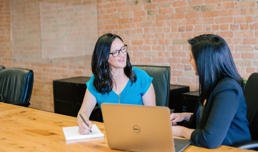 Two woman sitting together at a table talking, one writing on a pad of paper and the other next to a laptop