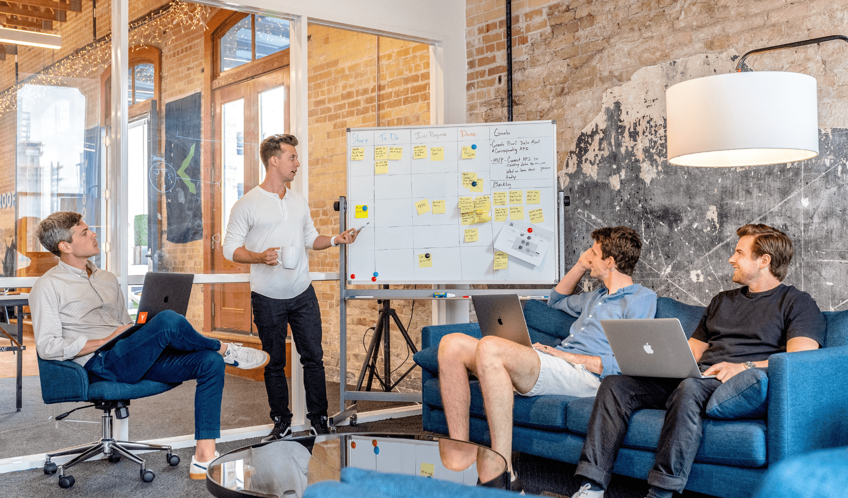 A group of people in a meeting room discuss a whiteboard, while one man leads the discussion