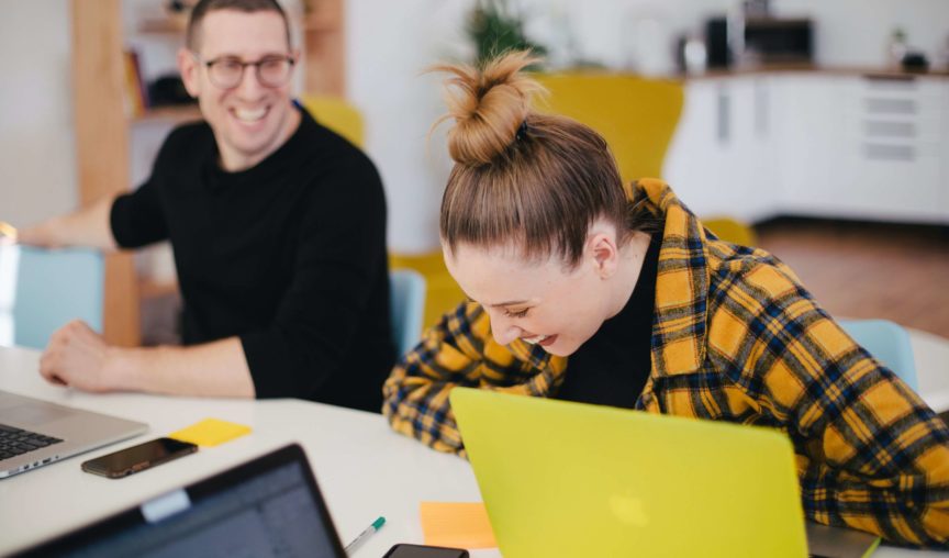 A male and female colleague seated laughing over a desk