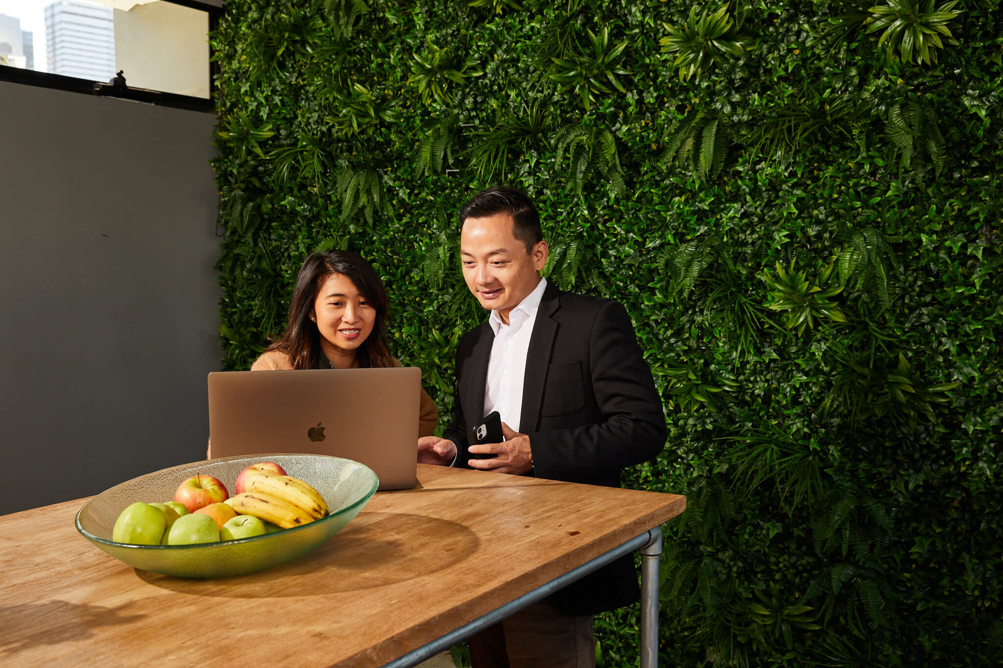 Two people standing in front of a leaf covered wall, looking at a laptop