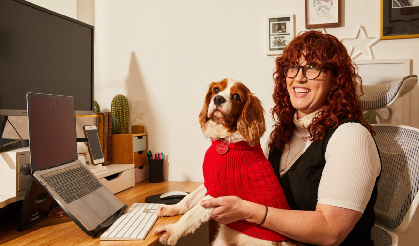 Woman sitting at home desk holding a dog in a red vest